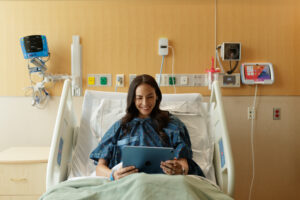 Patient resting in a hospital bed with HatchMed HallMonitor digital door signage displayed outside the patient room during a hospital renovation project.