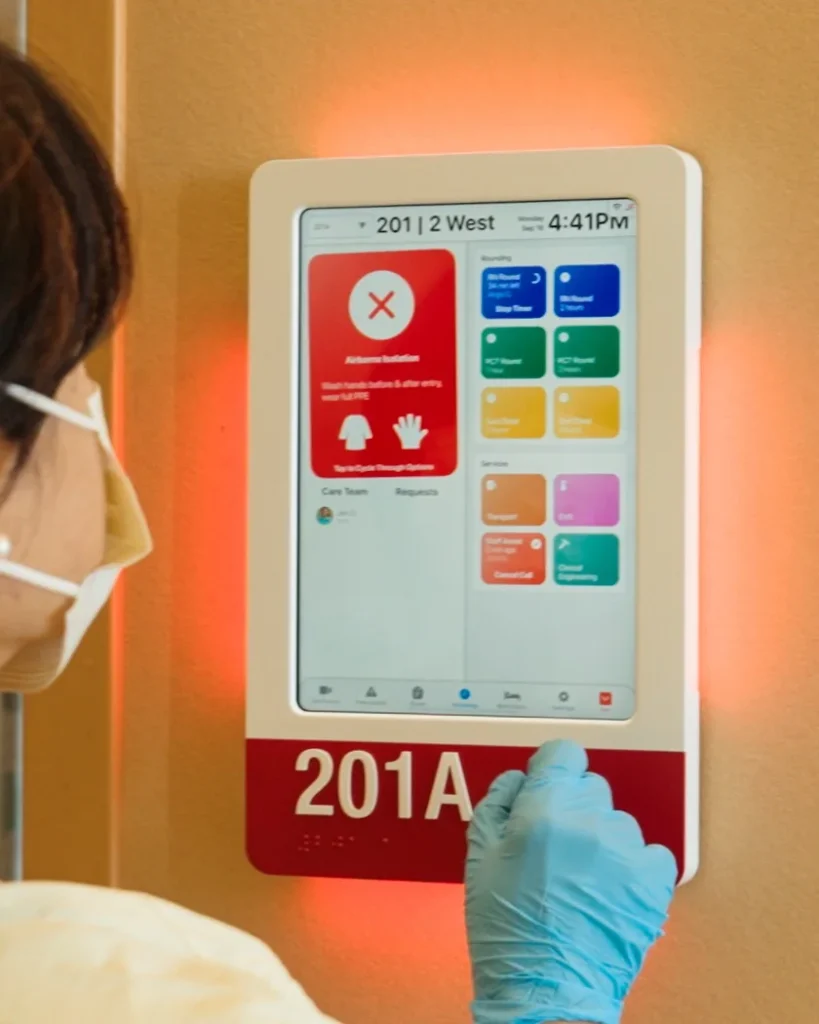 A nurse in an isolation gown and gloves taps a workflow button on a HallMonitor lit red for isolation.