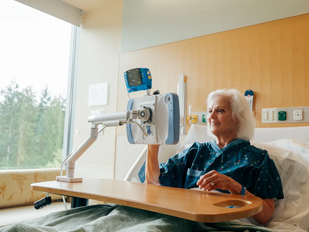 A PillowCase on a white arm clamped to an overbed table, beside a smiling patient by a window.