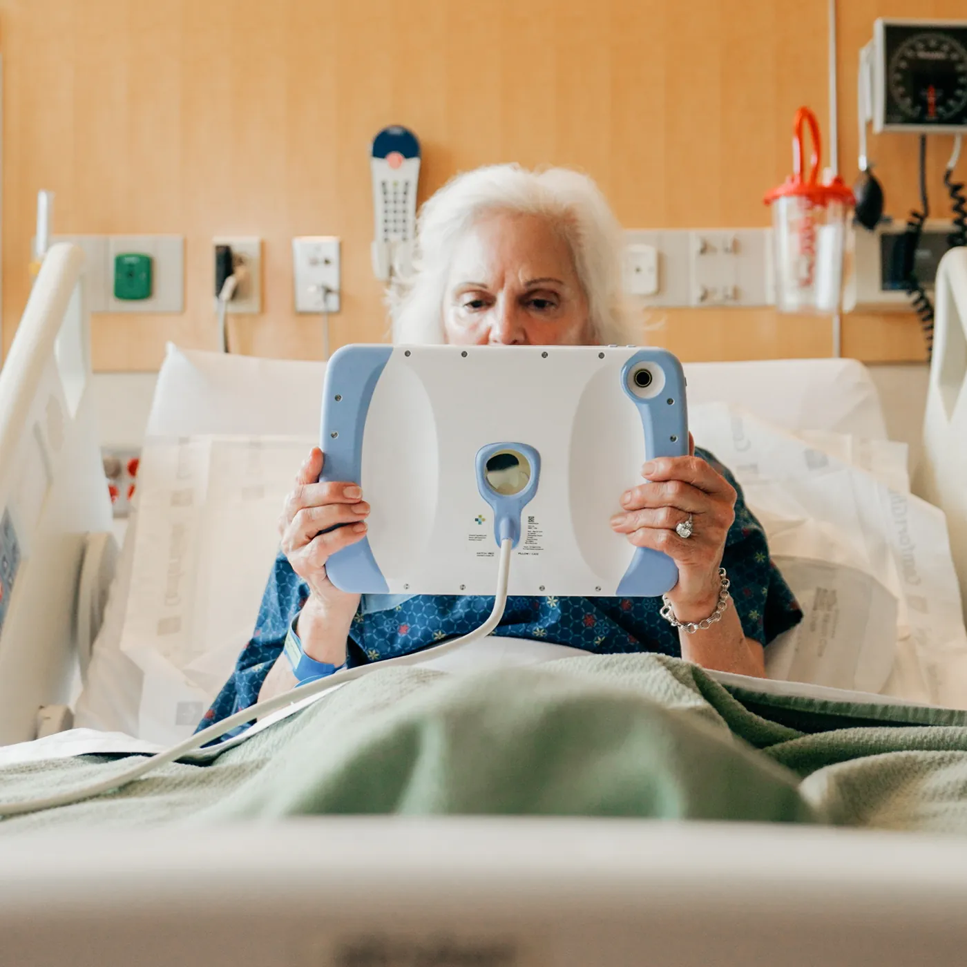 An older patient sitting up in bed holds a PillowCase in both hands, its cable trailing to the side.