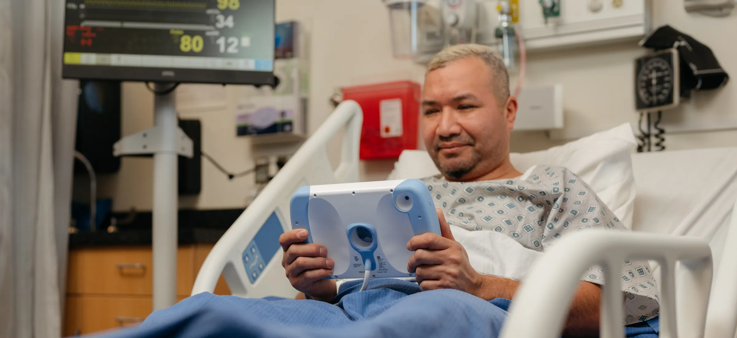 A patient sitting up in a hospital bed holds a PillowCase in both hands, its cable running down to the bed.